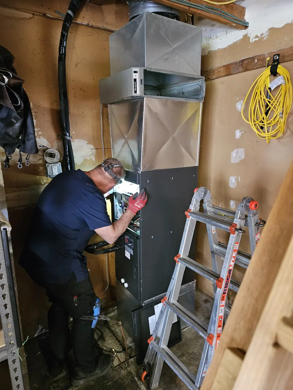 A technician wearing gloves and a headlamp working inside a residential furnace
