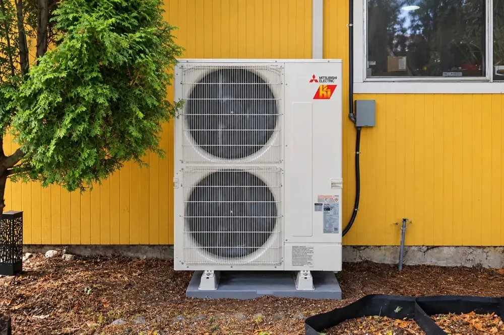 Mitsubishi outdoor heat-pump unit beside yellow house siding