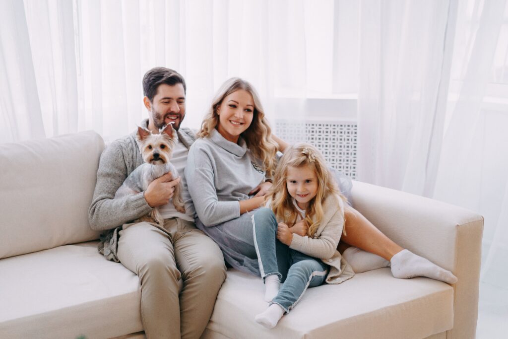 Happy family of four, including a mother, father, daughter, and small dog, sitting together on a cozy sofa in a bright living room, conveying warmth and comfort.