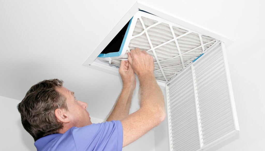 Man replacing an HVAC air filter in a ceiling vent, demonstrating air cleaning system maintenance for improved indoor air quality in Kent homes.
