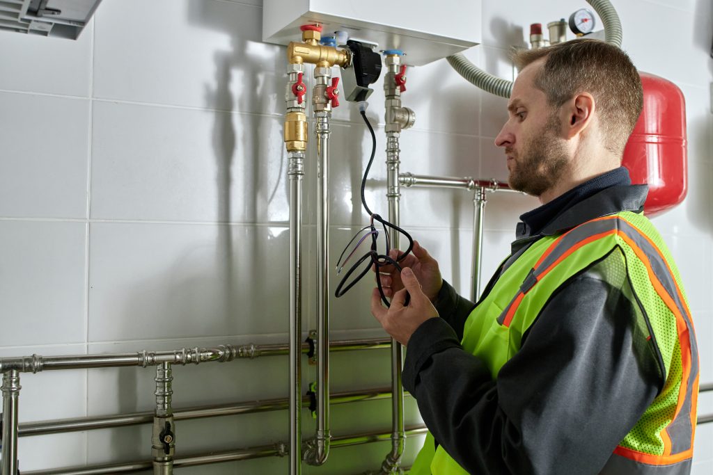 Caucasian middle-aged technician inspecting heating system components, including pipes and electrical connections, wearing safety gear in a residential setting.