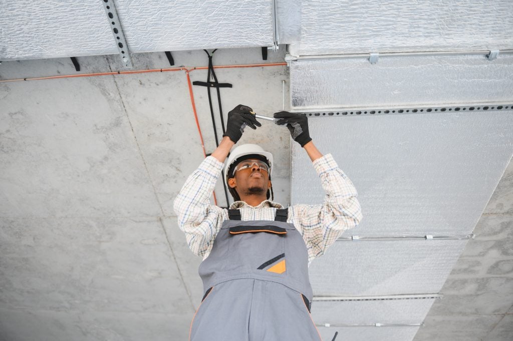 Construction worker installing ventilation system in a ceiling, wearing safety gear and gloves, representing smart electrical panel installation services for modern homes in Kent, WA.
