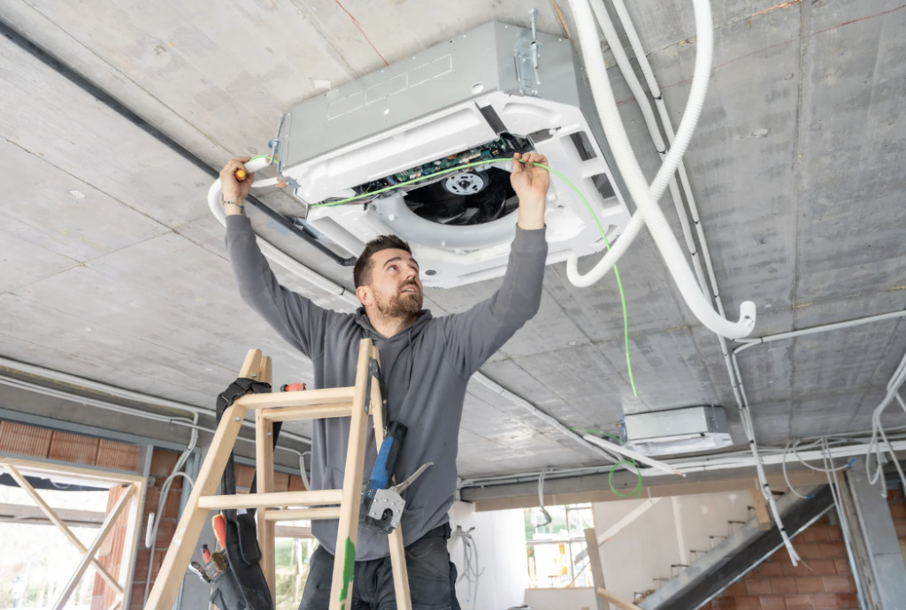 HVAC technician inspecting air duct system while standing on a ladder, emphasizing air duct cleaning and maintenance for improved indoor air quality before summer.